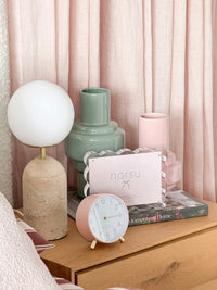 Maisie Silent Tick Alarm Clock in blush with gold accents on wooden bedside table next to decorative vases and books.