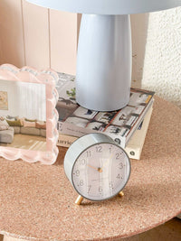 Maisie Silent Tick Alarm Clock in grey with gold accents on a bedside table next to a lamp and photo frame