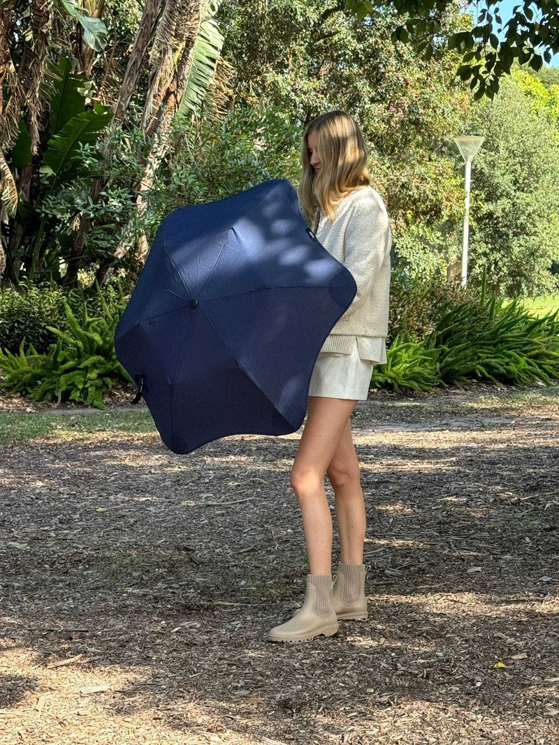 Woman holding a compact BLUNT Metro Umbrella in midnight navy outdoors among trees and greenery.