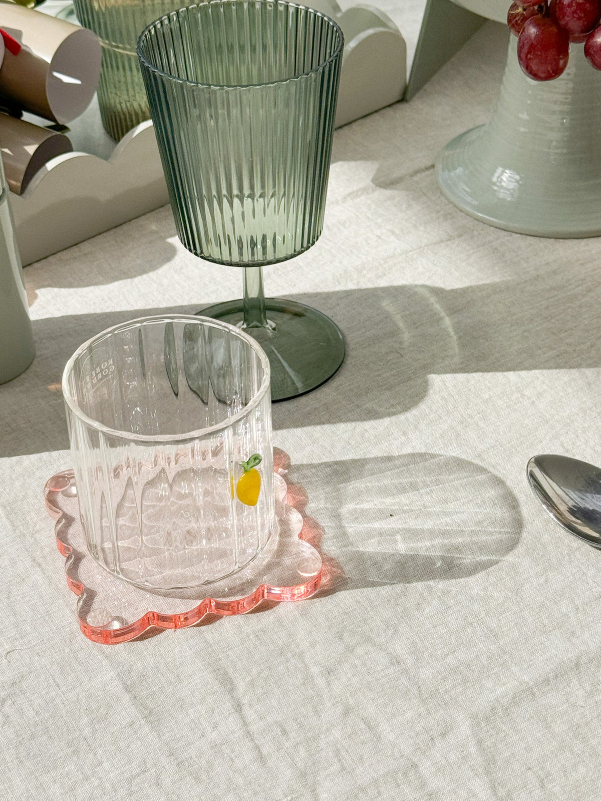 Green ribbed Saltara RPET tumbler and clear glass tumbler on white tablecloth with pink coaster and grapes nearby