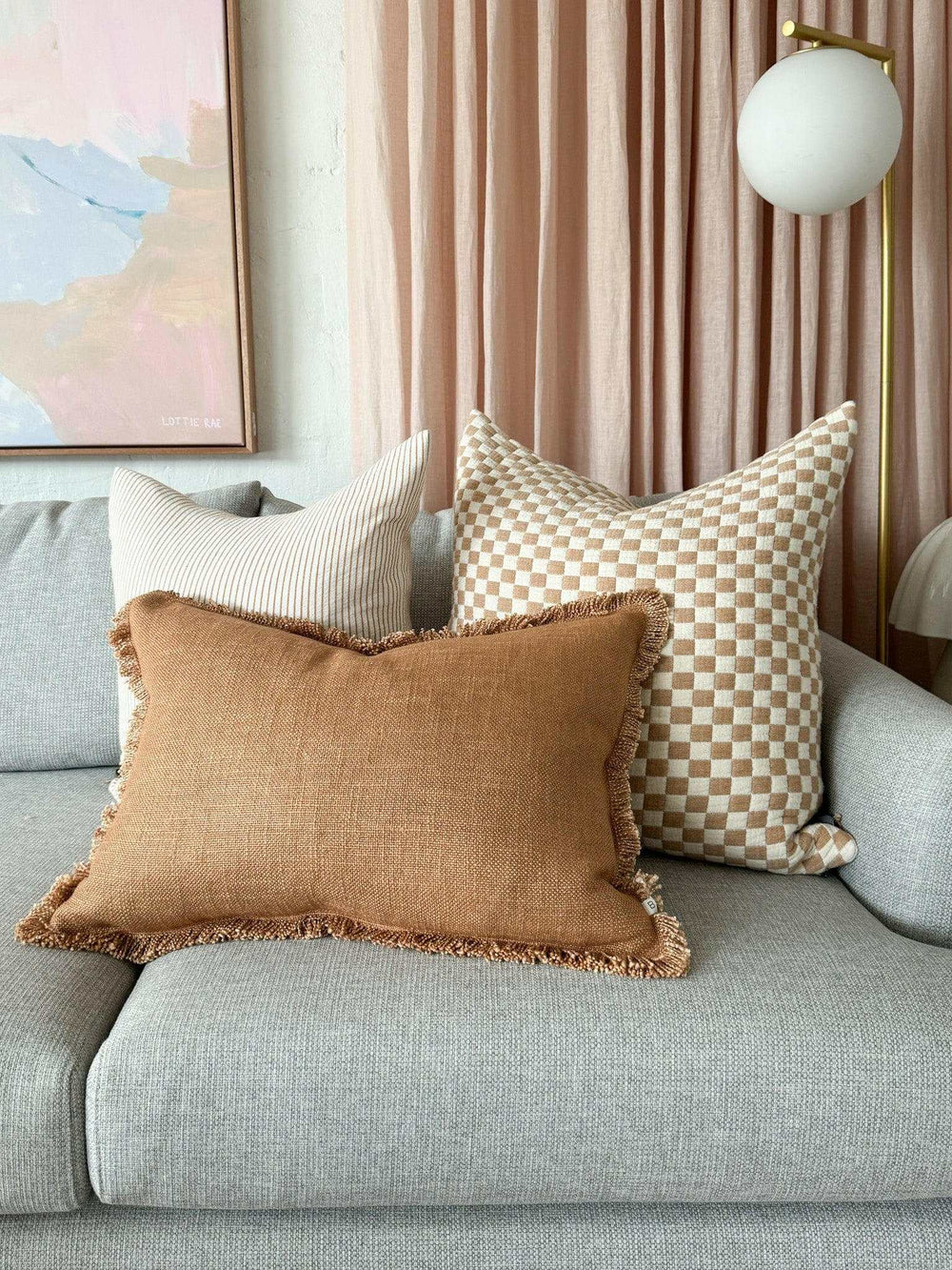 Baya Dover Cushion in nude with fringed edges on a gray sofa with decorative pillows and pink curtains background