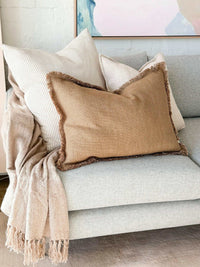 Baya Dover Cushion in nude with soft fringed edges on a light grey sofa with cozy throw and pillows