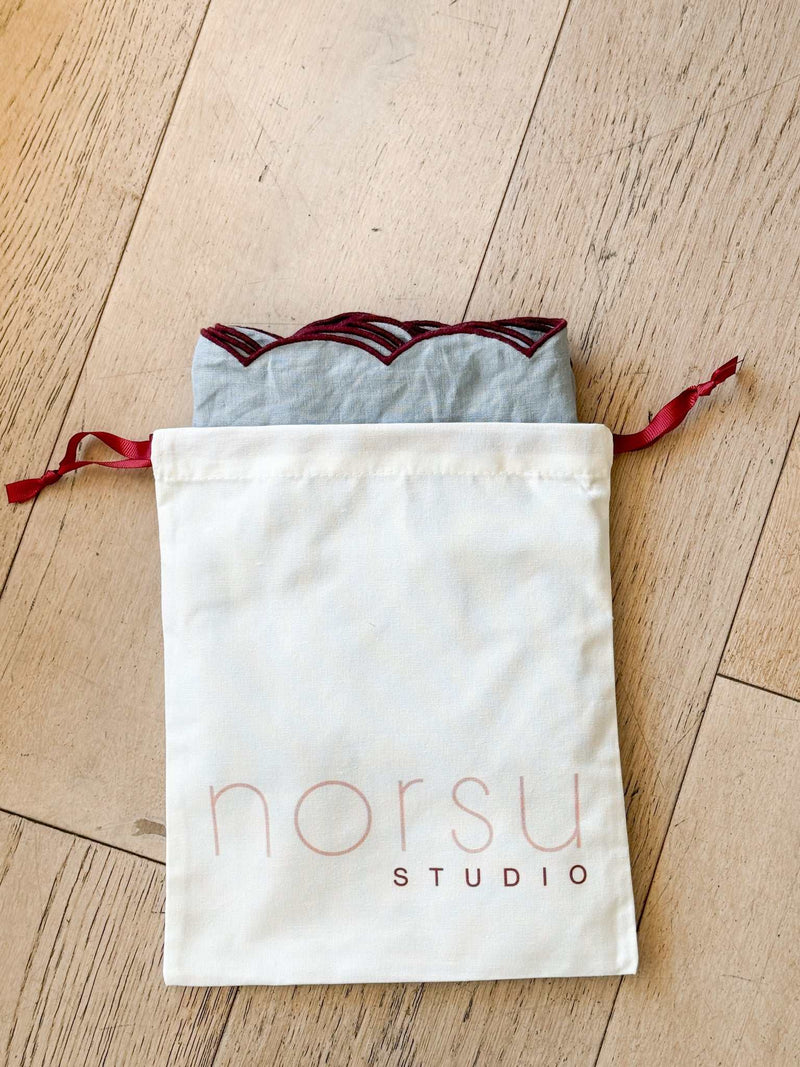 norsu Studio Linen scallop-edged blue napkins with deep burgundy edge in drawstring storage bag on wooden floor