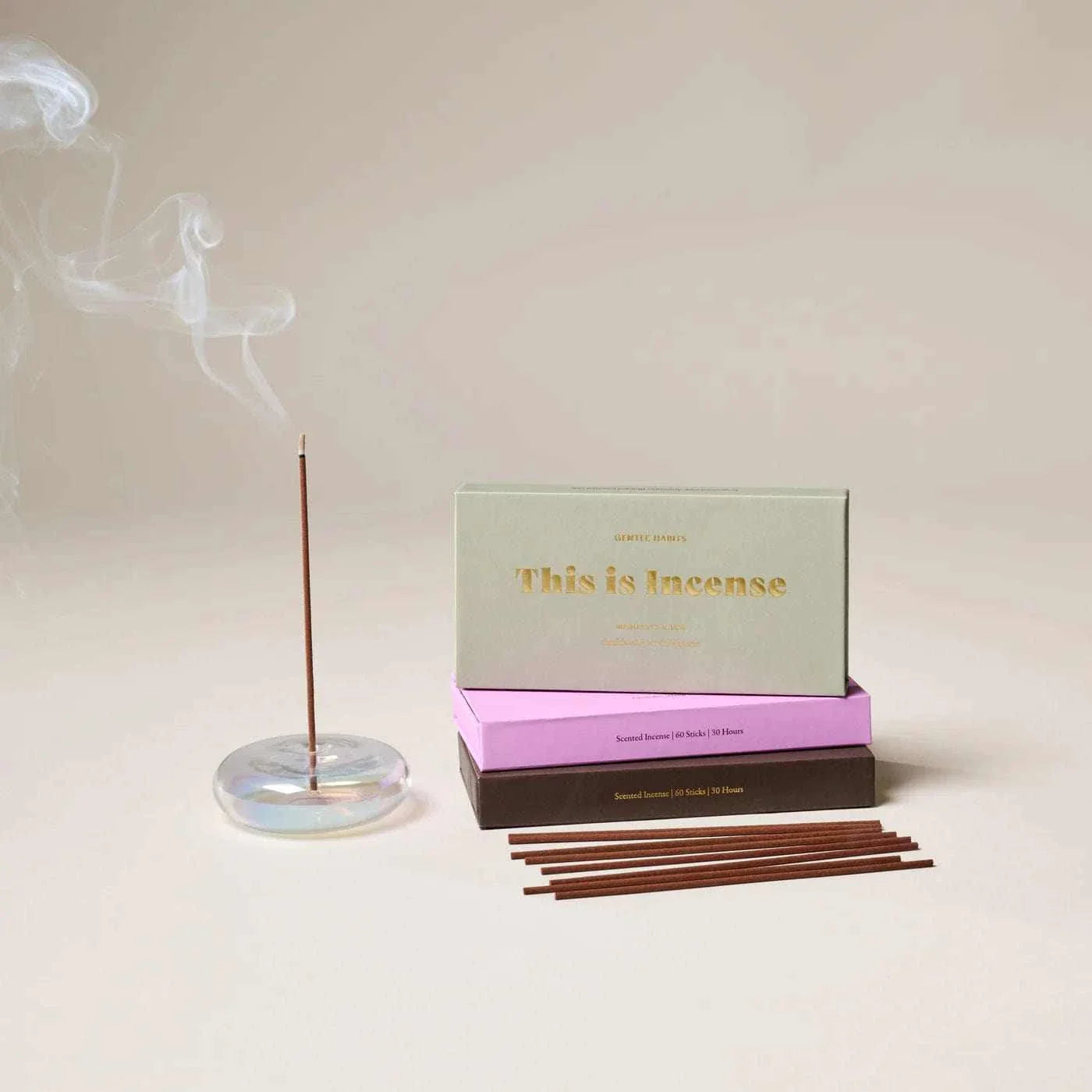 Gentle Habits Incense sticks burning with smoke in clear holder beside stacked pastel green, pink, and brown boxes on beige background