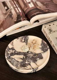 Round Linea marble tray in black and white Calacatta Viola pattern holding jewelry on dark surface with open book nearby