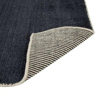 GlobeWest Tepih Neptune Rug in midnight color with hand-woven wool blend and subtle rustic texture, showing front and folded corner with striped backing.
