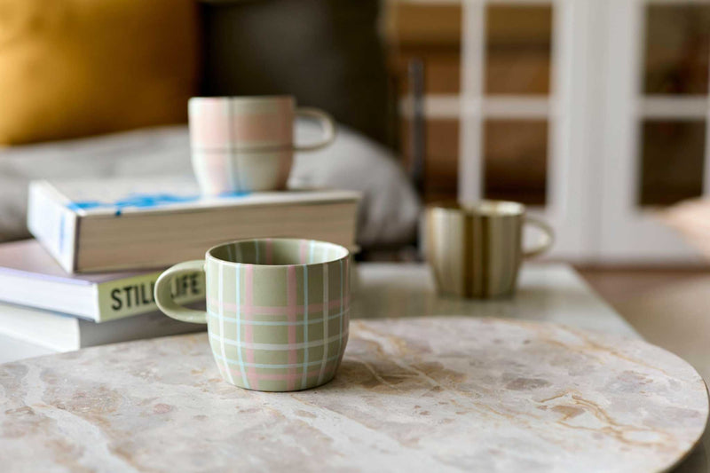 Robert Gordon hand-painted ceramic mug with olive tartan stripes on a marble table, surrounded by books and other patterned mugs