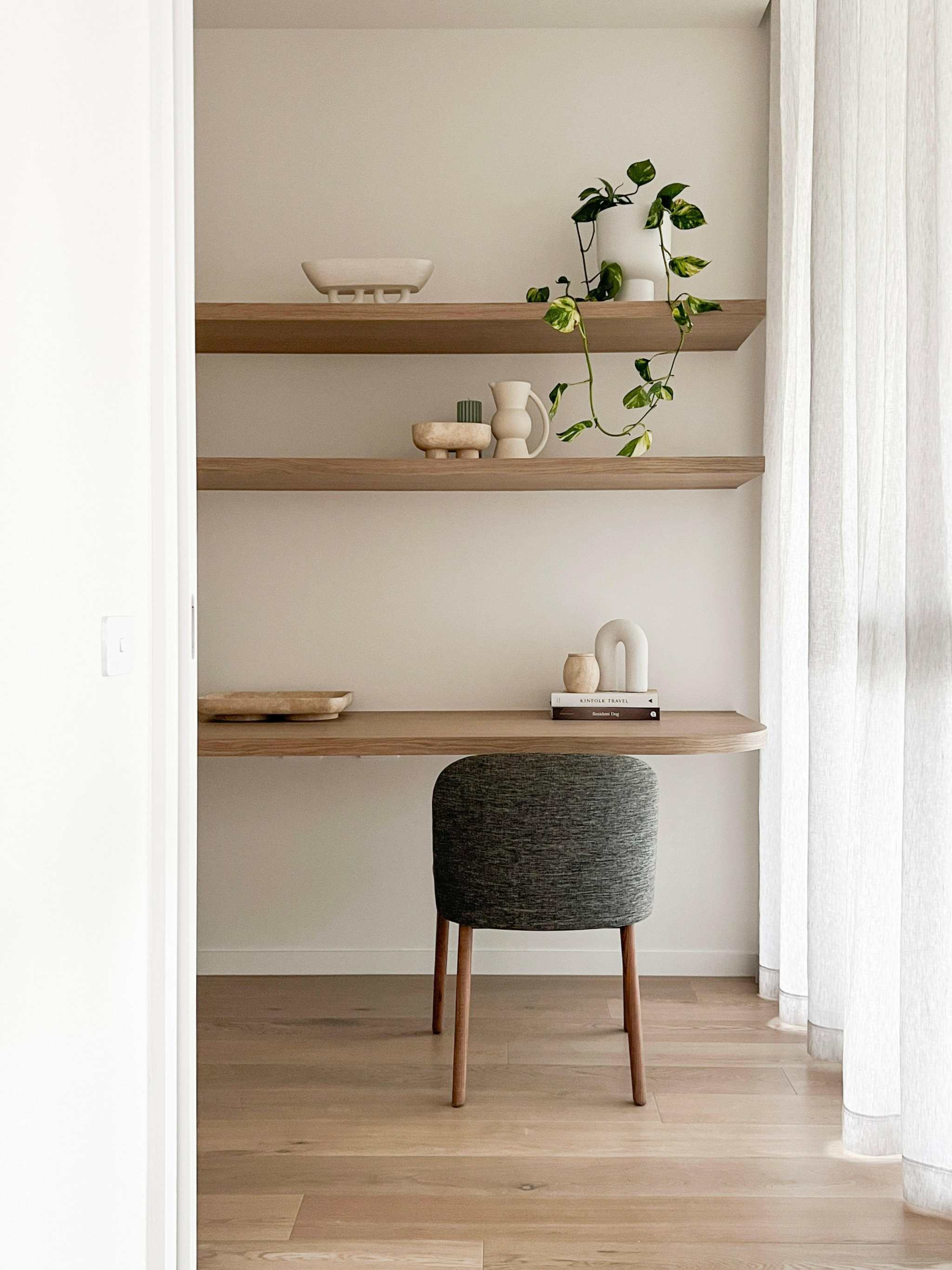 Minimalist home office with styled wooden shelves featuring decorative vases and a trailing plant, accompanied by a grey upholstered chair and sheer white curtains.