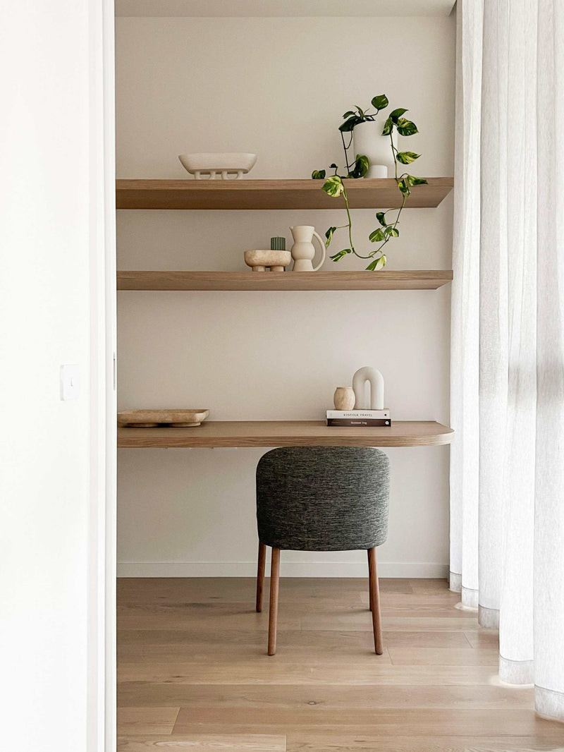 Minimalist home office with styled wooden shelves featuring decorative vases and a trailing plant, accompanied by a grey upholstered chair and sheer white curtains.