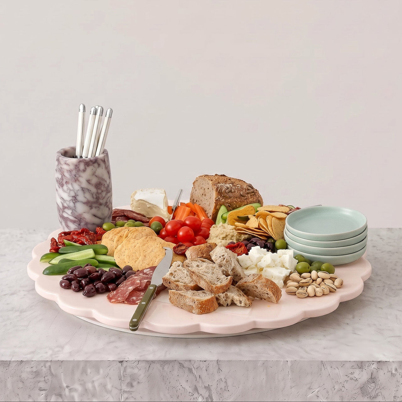 A pastel pink scalloped charcuterie board set on a light gray marble surface against a neutral light gray background. The board is styled with an assortment of food items including sliced bread, cheese, grapes, olives, crackers, nuts, and a marble cup holding metal utensils.