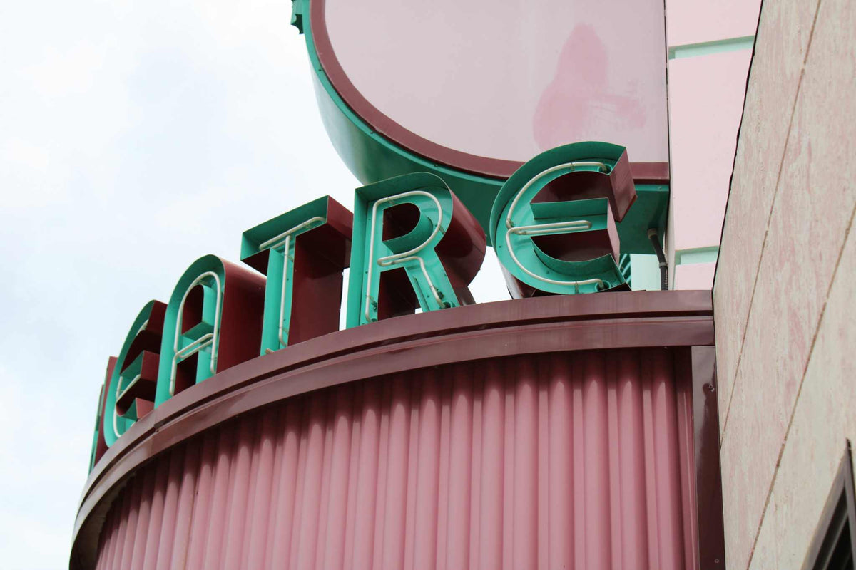 Vintage theatre sign with green neon letters on a maroon corrugated facade against a cloudy sky, theatre print wall art.