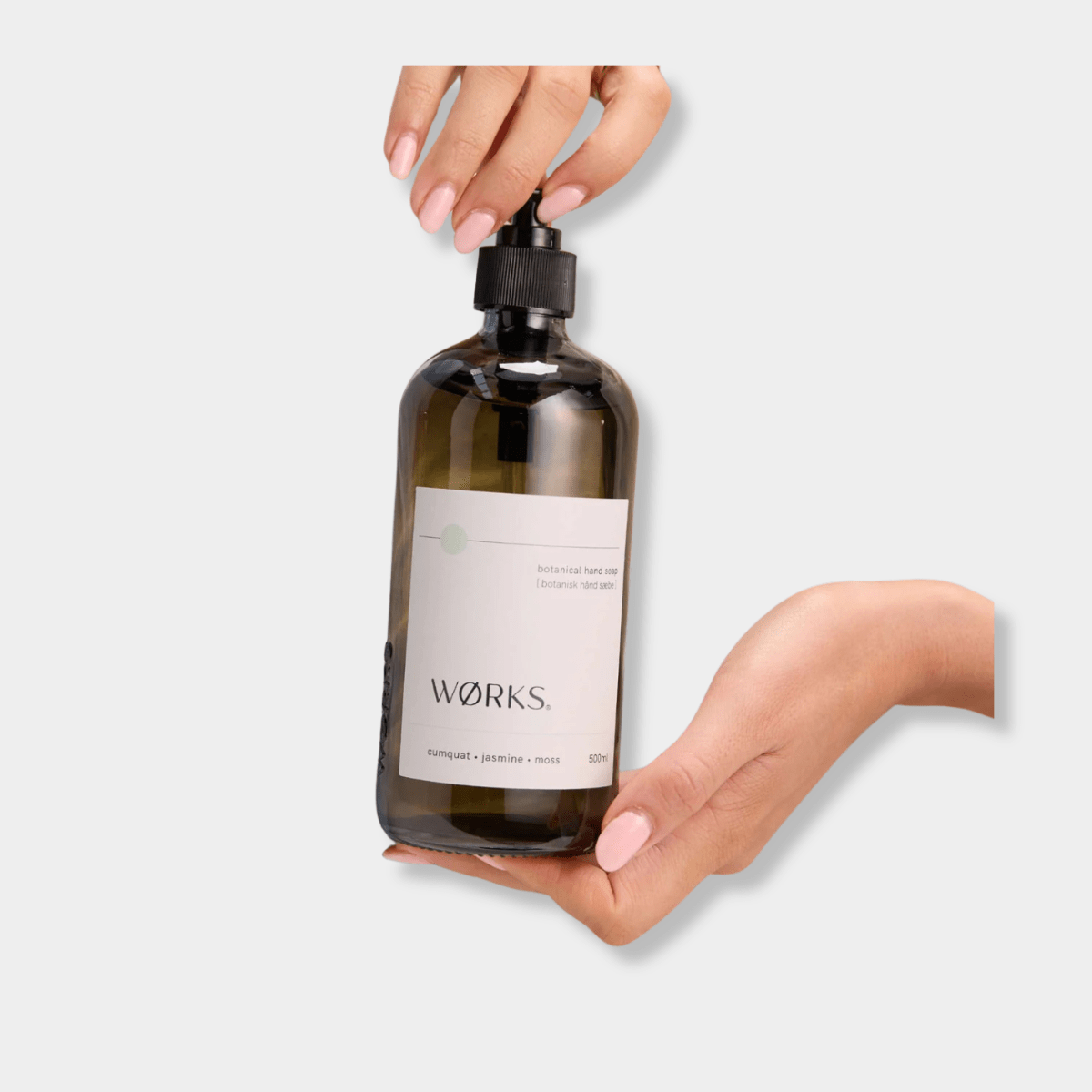 Works Hand Soap in smoky grey glass bottle with pump dispenser, cumquat and jasmine scent, held by hands with soft pink nails