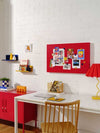 Red magnetic Story Board by Mustard Made with photos and notes, mounted on white brick wall above white desk and wooden chair, styled with red cabinet and colorful desk accessories matching Mustard Made lockers.