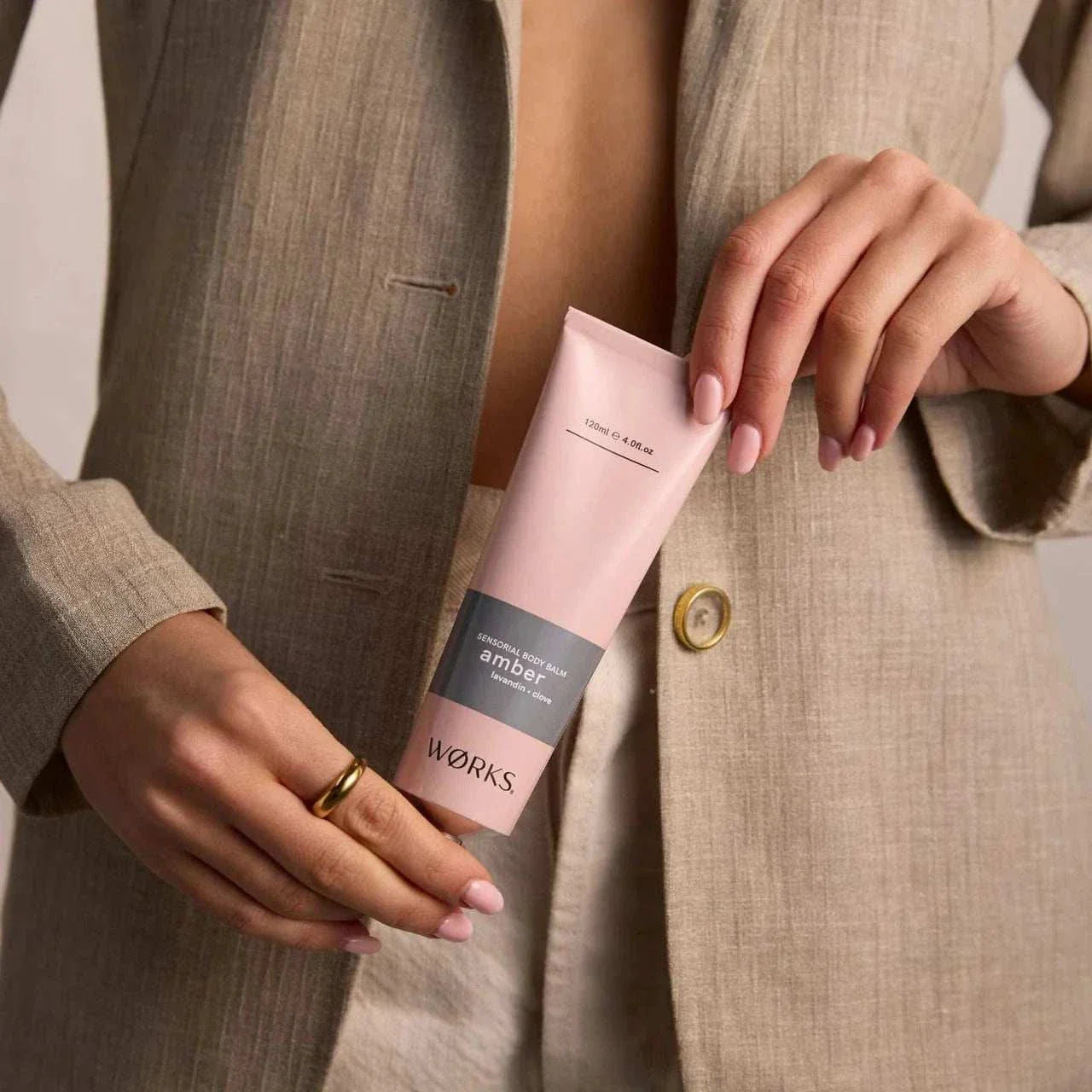 Works Body Balm tube in amber scent held by person wearing beige linen blazer
