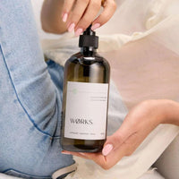 Works Hand Soap cumquat in a refillable smoky grey glass bottle with a pump dispenser held by a person with manicured nails