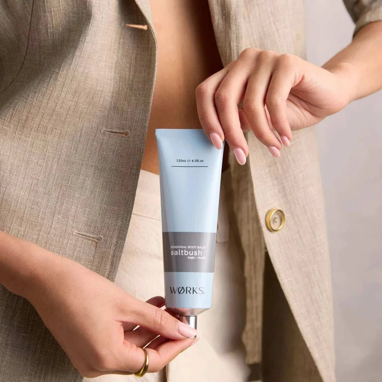 Works Body Balm tube in light blue with aluminium cap held by person wearing beige jacket, moisturizing skin with saltbush scent.