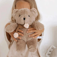 Soft caramel plush Prudence Bear with knitted chocolate scarf and pink pom poms held by child