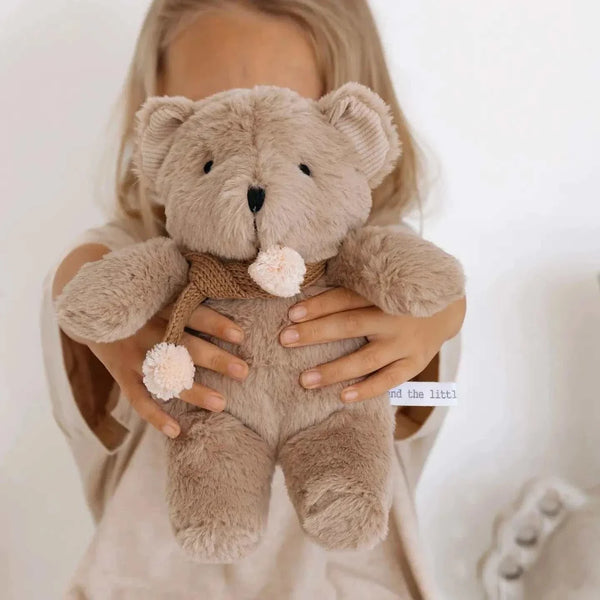 Soft caramel plush Prudence Bear with knitted chocolate scarf and pink pom poms held by child