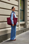 Woman carrying a burgundy Base Supply Take It Base Tote Bag with padded fabric and cotton shoulder straps outdoors.