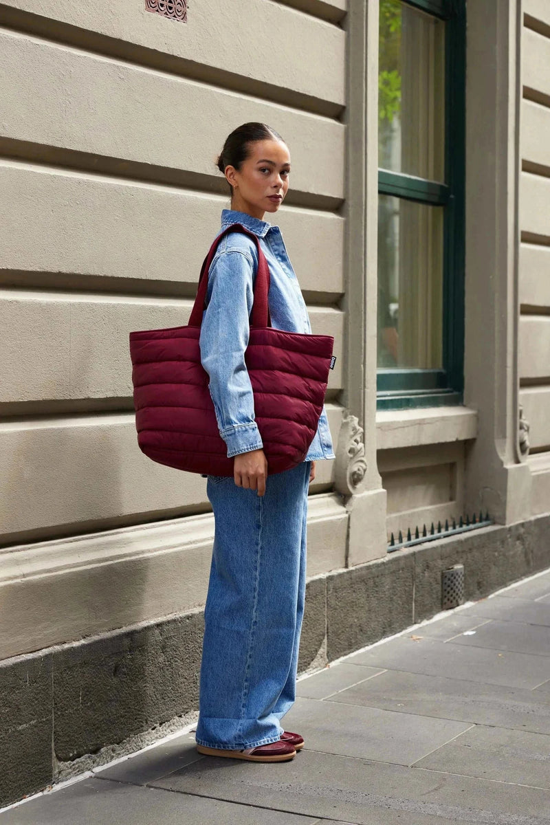 Woman carrying a burgundy Base Supply Take It Base Tote Bag with padded fabric and cotton shoulder straps outdoors.