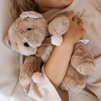 Caramel plush Prudence Bear with knitted chocolate merino wool scarf and pink pom poms, 25cm tall, cuddled by child.
