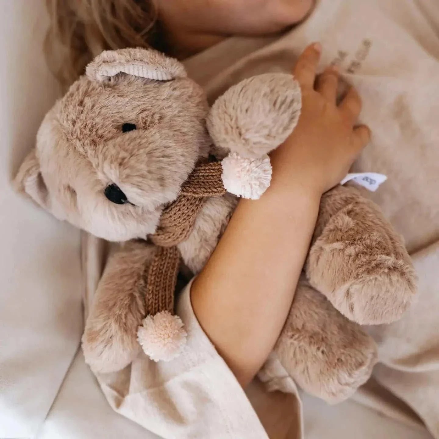 Caramel plush Prudence Bear with knitted chocolate merino wool scarf and pink pom poms, 25cm tall, cuddled by child.