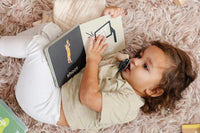 Baby lying on soft rug holding and looking at a basketball-themed board book titled "Basketball Baby"