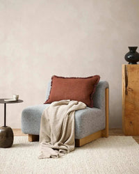 Baya Dover Cushion in brick color with soft fringed edges on a modern grey armchair with beige throw in a minimalist living room