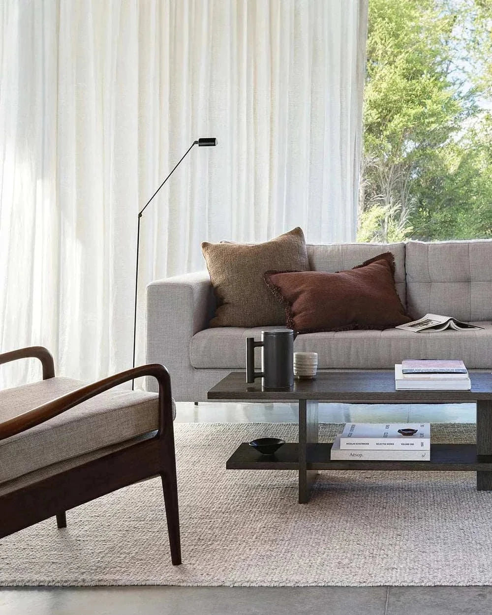 Living room with grey sofa featuring Baya Dover Cushion in brick color and brown cushions, modern wooden armchair, and coffee table with books and decor