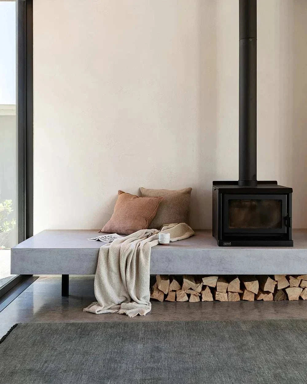 Eadie Flaxmill Cushion in clay color on a minimalist concrete bench next to a black wood stove with firewood storage underneath.