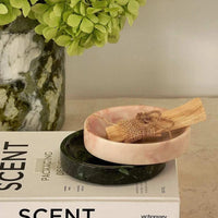 Black Blaze Linea small pink marble dish holding wooden sticks, stacked on a black marble dish on a table with a plant and a book nearby