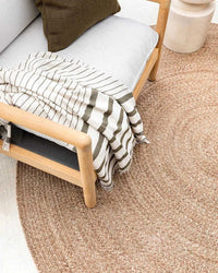 Round Baya Mornington Floor Rug in teak color, handbraided flatweave made from recycled PET fiber, 210cm diameter, natural earthy tones placed under a wooden chair with a striped throw blanket.