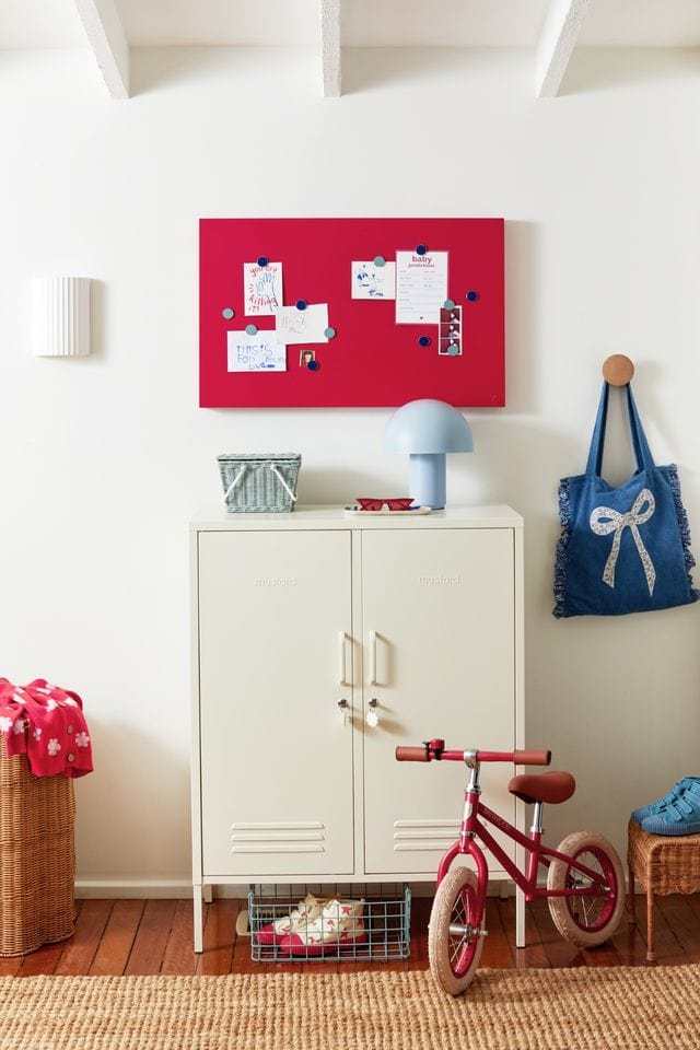 Red magnetic Storyboard with notes, mounted above cream Mustard Made lockers in a bright, organized room with children's bike and accessories.