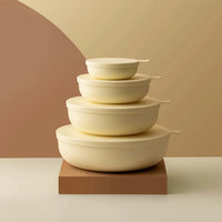 Set of four cream-colored Styleware nesting bowls with lids stacked on a brown platform against a beige and brown background