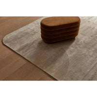 GlobeWest Tepih Curve Rug in sand color with subtle curved pattern on wooden floor under brown ottoman