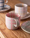 Robert Gordon Hand-Painted Mug with pink tartan checks, ceramic stoneware, 350ml capacity on wooden table with matching plates and another mug in background