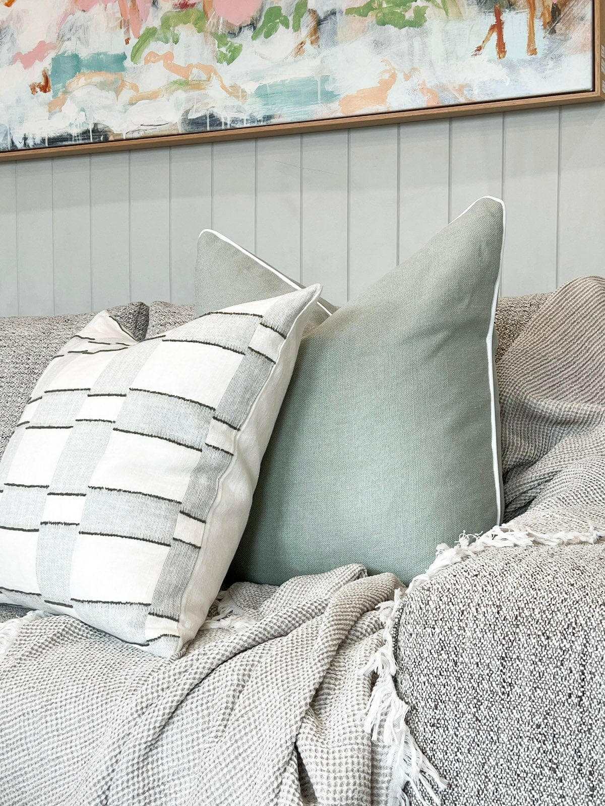 norsu Studio Cushion in Haven Celadon with white leather piping on a textured gray sofa with a neutral patterned cushion and a soft throw blanket