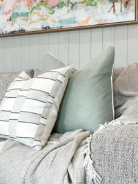 norsu Studio Cushion in Haven Celadon with white leather piping on a textured gray sofa with a neutral patterned cushion and a soft throw blanket