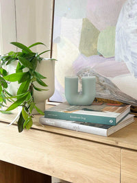 Black Blaze Curl Curl sculptural candle on stack of books beside green leafy plant on wooden surface.