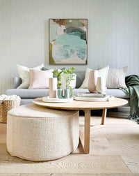 norsu Design Your Own Ottoman in soft boucle fabric with blush leather piping, placed next to a round wooden coffee table in a modern living room setting