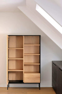 Ethnicraft Blackbird Storage Cupboard in natural oak with black frame, featuring shelves and drawer, standing in a modern room corner