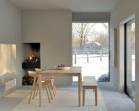 Ethnicraft Bok bench seat in natural oak placed in a modern dining room with a large window and fireplace.