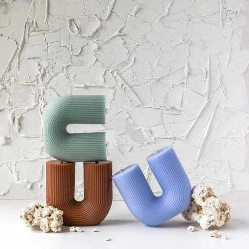 Three sculptural Black Blaze candles in sage green, brown, and blue colors with a wavy curled design stacked against a textured white wall background.