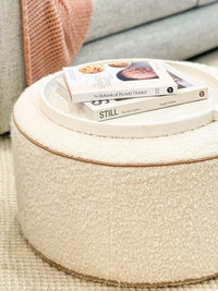 Round soft upholstered ottoman with blush leather piping and woolen fabric, styled with a white tray and stacked books on top.