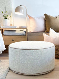 Customizable round white boucle fabric ottoman with blush leather piping in a cozy living room setting