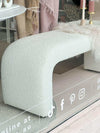 Curved oatmeal Zaru Boucle Bench upholstered with textured boucle fabric, placed indoors near window display