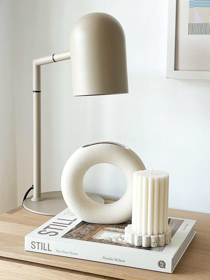 Pia Task Lamp in bone color with modern architectural design on a wooden desk next to decorative objects and books
