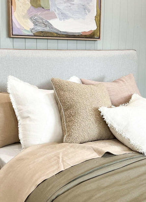 norsu Studio Bouclé Buff cushion with white leather piping on bed with neutral throw pillows and soft bedding