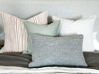 norsu Studio Cushion in celadon green with white leather piping, displayed with neutral striped and textured cushions on a bed
