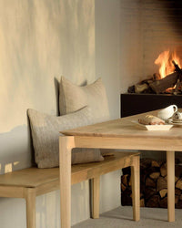 Ethnicraft Bok bench seat in natural oak wood beside a dining table with cushions and a fireplace in the background